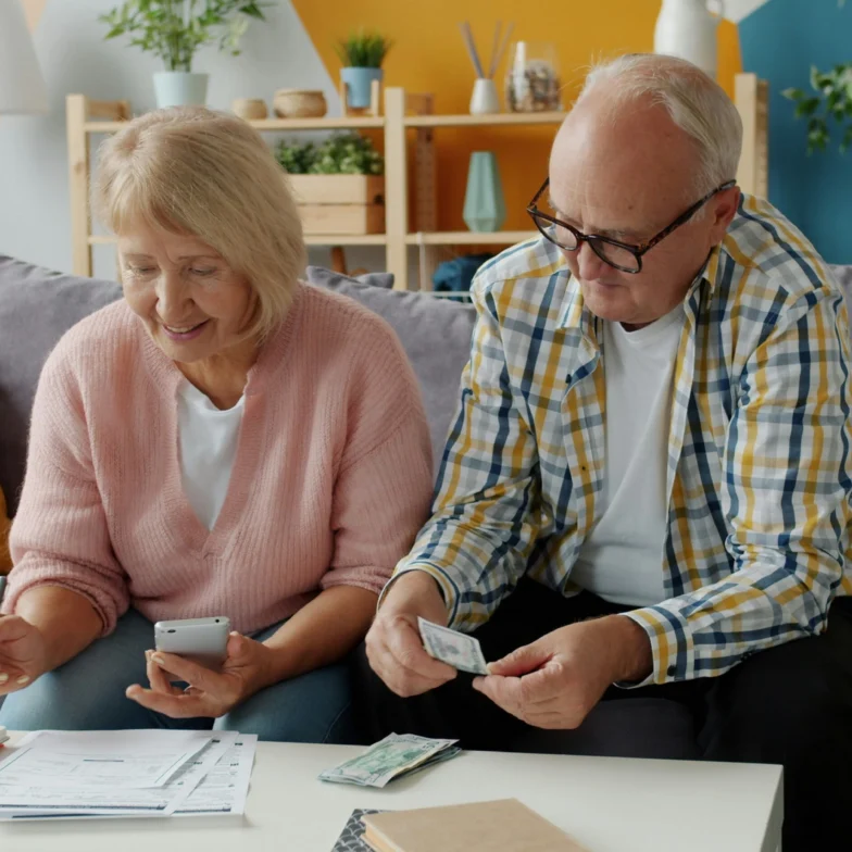 An older woman and man sit on a couch, reviewing paperwork and counting cash at a coffee table in a living room.