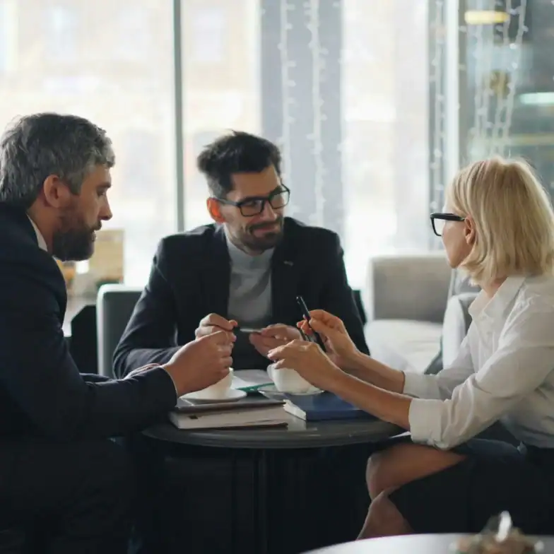 Three people in business attire sit at a table in an office, having a discussion with documents and notebooks in front of them.