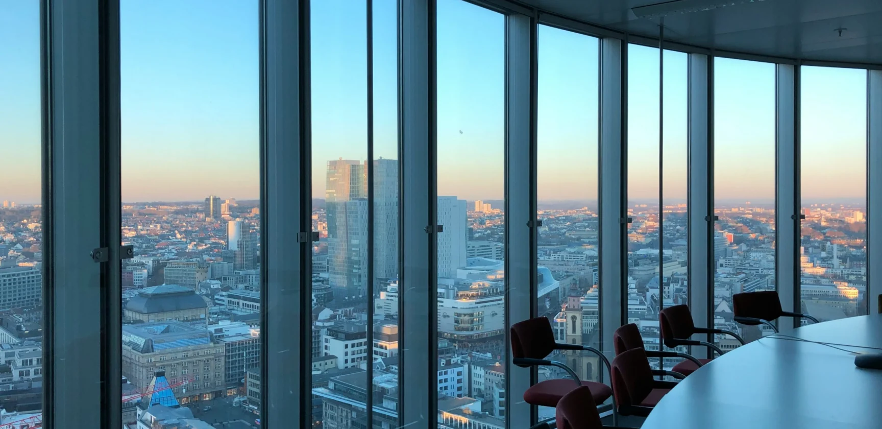 A modern conference room with red chairs and a round table overlooks a city skyline through floor-to-ceiling windows at sunset.