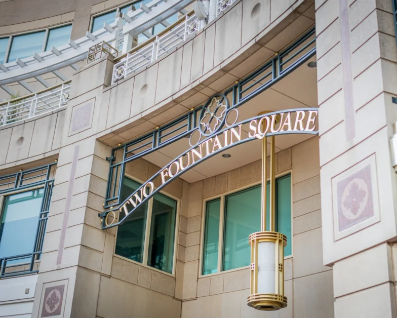 Sign reading “Two Fountain Square” hangs on a modern building facade with large windows, decorative railings, and a cylindrical hanging light fixture.