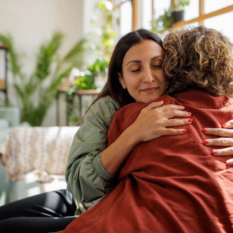 Two women are sitting indoors and hugging each other closely. One woman faces the camera with her eyes closed, while the other has her back to the camera.