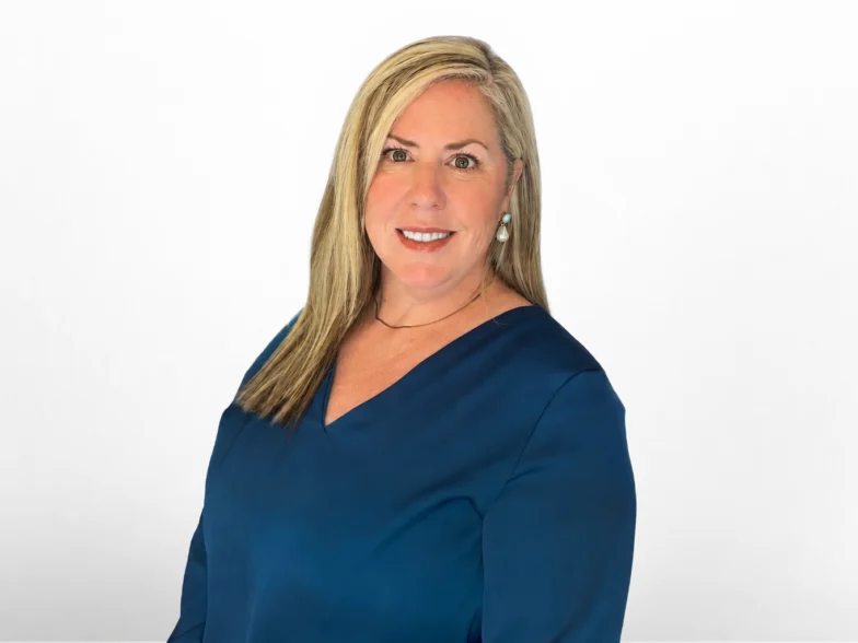 A professional headshot of a woman with long blonde hair wearing a blue v-neck top.